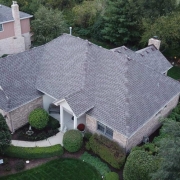 Aerial view of a suburban house with a grey residential roofing, surrounded by well-maintained landscaping, green lawns, and mature trees. A driveway leads to the garage at the front of the house.
