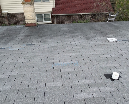 A newly installed gray asphalt shingle roof, indicative of a recent roof replacement, features several vents and black patches, with a house in the background.