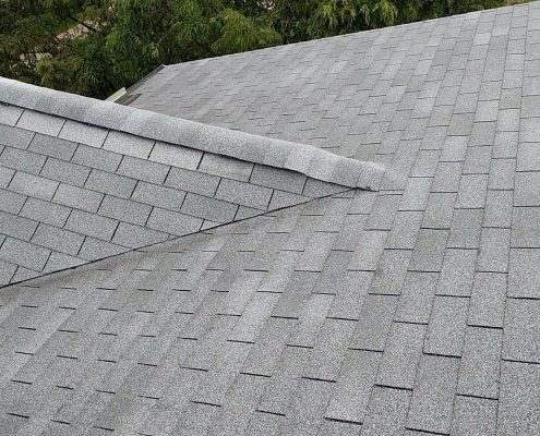 An image of a roof covered with gray asphalt shingles, showcasing the intersection of two roof slopes with a central ridge vent. This typical scene from a professional roof installation has trees visible in the background.