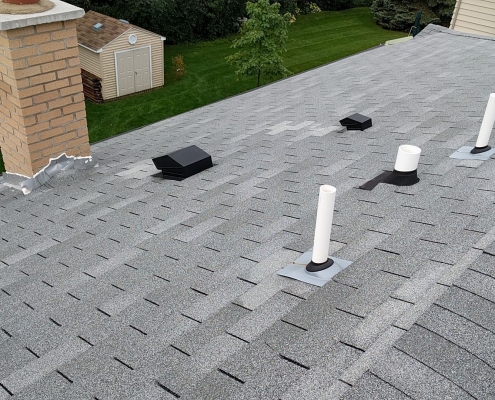 A rooftop with grey shingles featuring multiple vent pipes and a chimney suggests the work of a skilled roofing contractor. A lush green yard with a shed and trees is visible in the background, completing the picturesque residential roofing scene.
