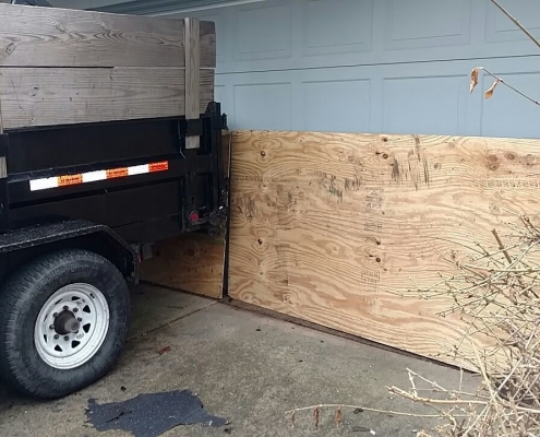 A trailer backs into a driveway, placing a large wooden board against a closed garage door, indicating preparations for roof installation.