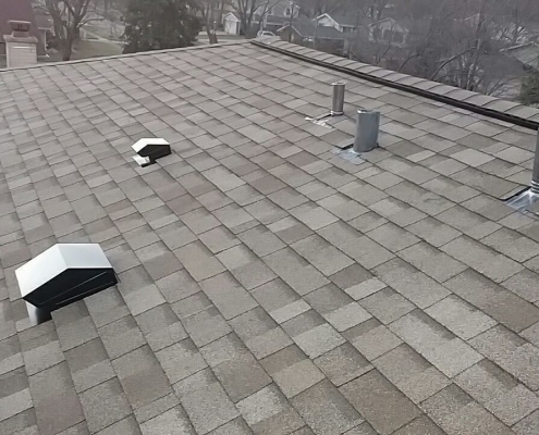 A view of a residential rooftop with gray shingles, showcasing expert residential roofing, featuring multiple vents and chimneys. Trees and neighboring roofs are visible in the background.