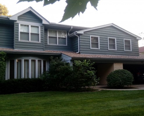 A two-story house with green siding, a double garage, and a well-maintained lawn and shrubs stands proudly. The pickup truck parked in the driveway hints at recent residential roofing work or perhaps an upcoming roof replacement.