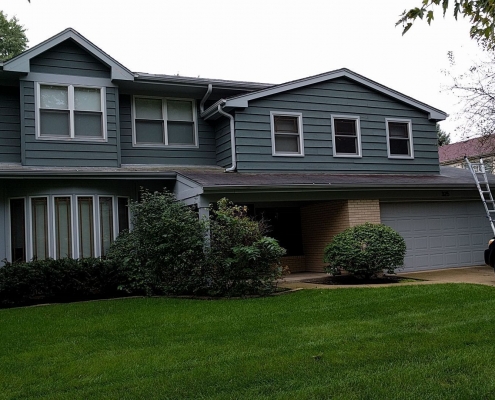 A two-story house with dark green siding, a garage, and a well-maintained lawn. A ladder leans against the right side near a black SUV parked in the driveway, indicating ongoing roof installation. Trees surround the property.