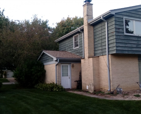A two-story house with green siding and a brick lower level, featuring a chimney, a lush lawn, and trees in the background. A small white door leads into an extension on the side of the house. The roofing contractor recently completed an efficient roof replacement, adding to its charm.