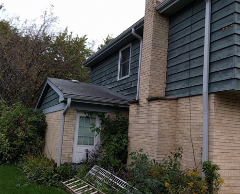 A suburban house with gray siding and beige brick has a white trellis leaning against it surrounded by shrubs and trees. Recently, a roofing contractor completed a roof replacement that perfectly complements the home's aesthetic.