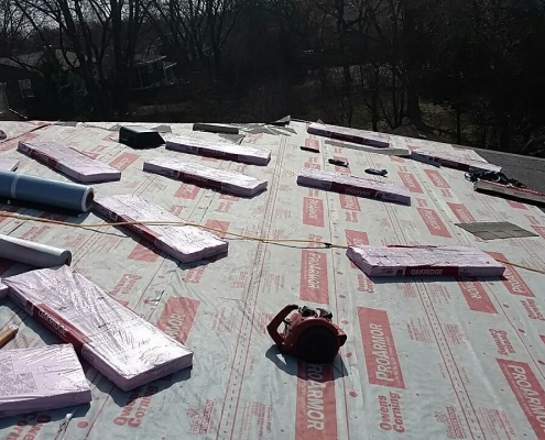 A residential roofing project is under construction with insulation material and tools scattered on the surface, including wrapped insulation sheets and a blower. Trees and rooftops are visible in the background, showcasing the meticulous work of a professional roofing company.