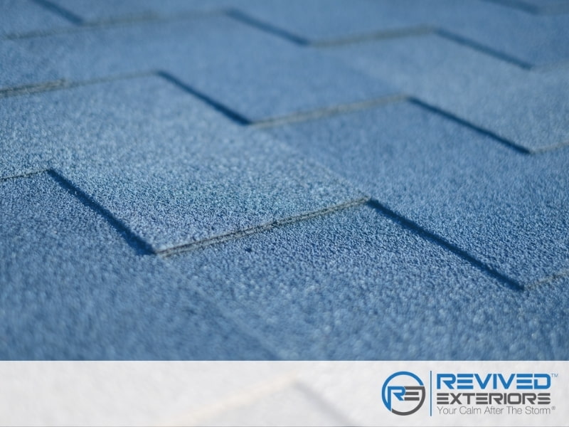 Close-up view of blue asphalt roof shingles with the "Revived Exteriors" logo in the lower right corner, showcasing our expertise as a leading roofing contractor. Close-up view of blue asphalt roof shingles with the "Revived Exteriors" logo in the lower right corner, showcasing our expertise as a leading roofing contractor.